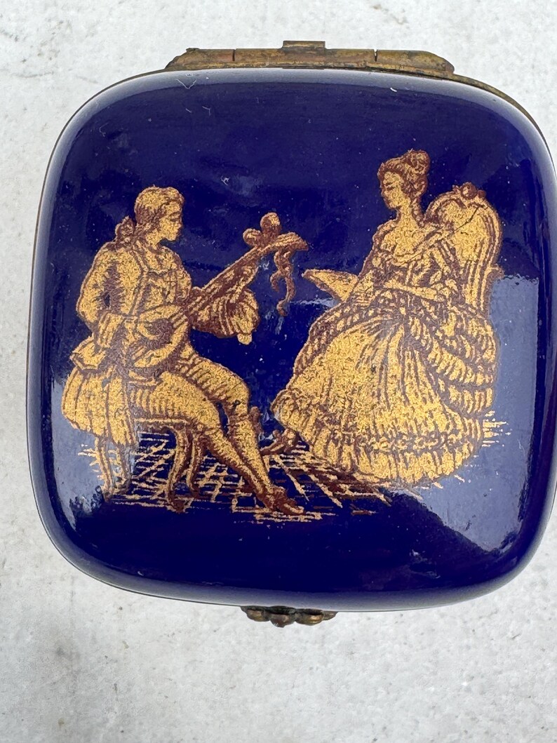 Puede incluir: Caja de baratijas rectangular azul marino con ribete met&aacute;lico dorado. La tapa presenta una ilustraci&oacute;n dorada de un hombre tocando un instrumento de cuerda y una mujer sentada en una silla.