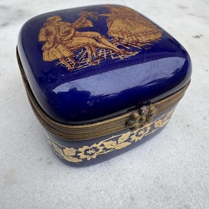 Puede incluir: Una caja de baratijas rectangular azul cobalto con un borde de metal dorado. La tapa presenta una ilustraci&oacute;n dorada de un hombre tocando un instrumento y una mujer. La base de la caja tiene un dise&ntilde;o floral dorado.