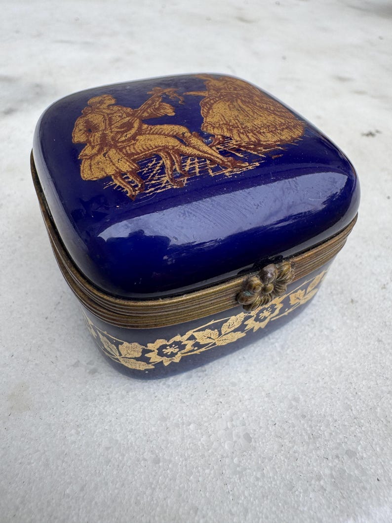 Puede incluir: Una caja de baratijas rectangular azul cobalto con un borde de metal dorado. La tapa presenta una ilustraci&oacute;n dorada de un hombre tocando un instrumento y una mujer. La base de la caja tiene un dise&ntilde;o floral dorado.