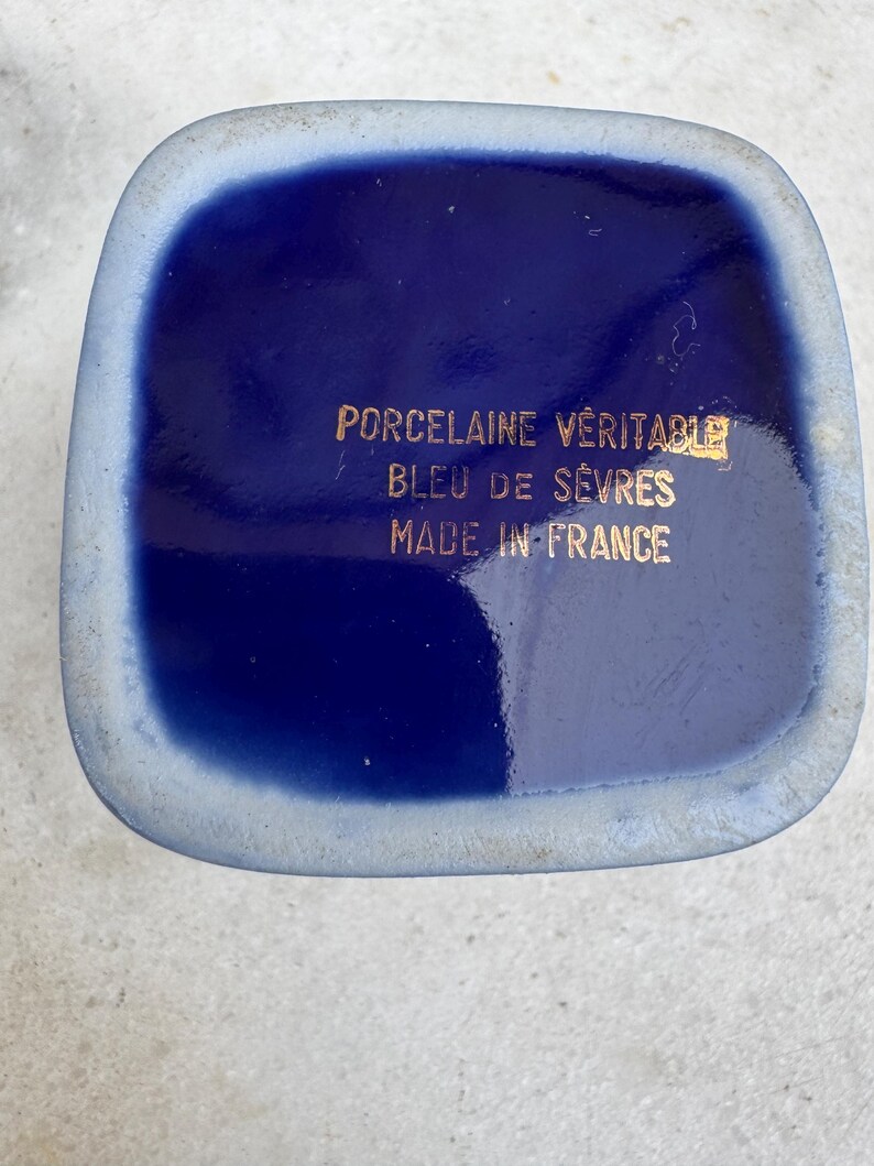 Puede incluir: Primer plano de un objeto cuadrado de porcelana azul cobalto con esquinas redondeadas. El texto dorado dice "PORCELAINE V&Eacute;RITABLE BLEU DE S&Egrave;VRES MADE IN FRANCE". El objeto tiene un borde azul claro.