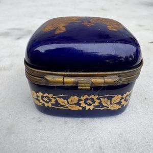 Puede incluir: Peque&ntilde;a caja de baratijas azul cobalto con un dise&ntilde;o floral dorado alrededor de la base y en la tapa. La caja tiene una tapa con bisagras y un marco de metal dorado. La caja es rectangular con bordes redondeados.