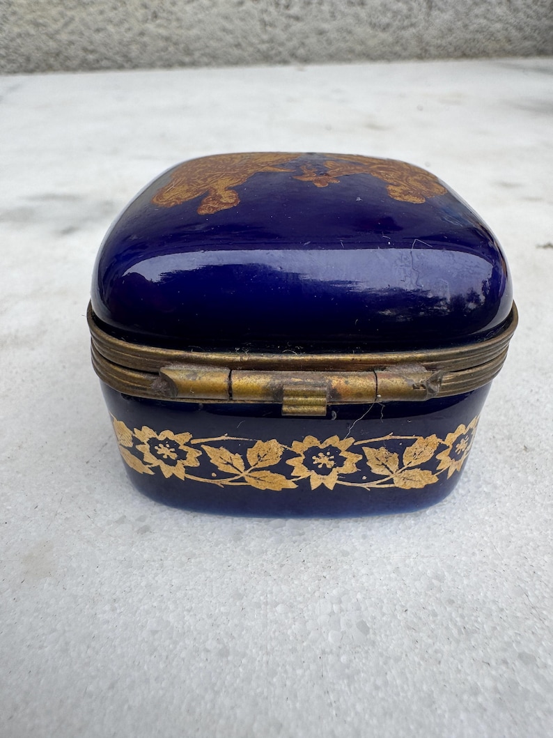 Puede incluir: Peque&ntilde;a caja de baratijas azul cobalto con un dise&ntilde;o floral dorado alrededor de la base y en la tapa. La caja tiene una tapa con bisagras y un marco de metal dorado. La caja es rectangular con bordes redondeados.