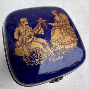 Puede incluir: Una caja de baratijas cuadrada de color azul cobalto con una ilustraci&oacute;n dorada de un m&uacute;sico y una mujer con un vestido. La caja tiene una tapa con bisagras y un borde de metal de color bronce.