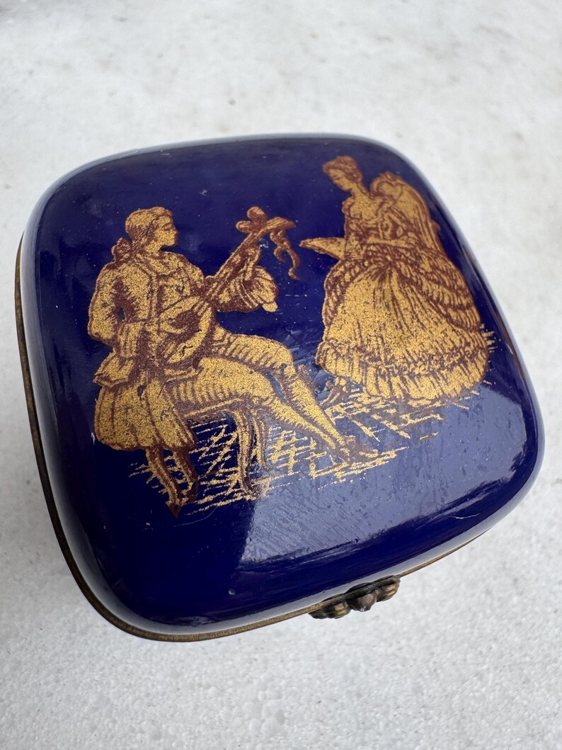 Puede incluir: Una caja de baratijas cuadrada de color azul cobalto con una ilustraci&oacute;n dorada de un m&uacute;sico y una mujer con un vestido. La caja tiene una tapa con bisagras y un borde de metal de color bronce.