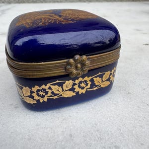 Puede incluir: Peque&ntilde;a caja de baratijas azul cobalto con un dise&ntilde;o floral dorado en la base y un &aacute;guila dorada en la tapa. La caja tiene una tapa con bisagras y un cierre decorativo. La caja probablemente est&aacute; hecha de porcelana o cer&aacute;mica.