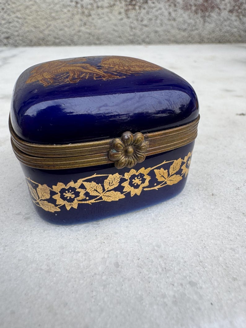 Puede incluir: Peque&ntilde;a caja de baratijas azul cobalto con un dise&ntilde;o floral dorado en la base y un &aacute;guila dorada en la tapa. La caja tiene una tapa con bisagras y un cierre decorativo. La caja probablemente est&aacute; hecha de porcelana o cer&aacute;mica.