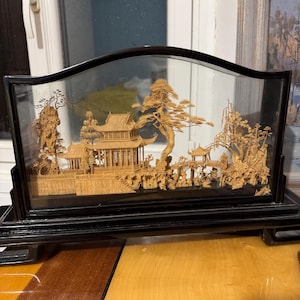 May include: A detailed wooden diorama of an Asian landscape scene, enclosed in a black framed display case. The scene features intricate carvings of buildings, trees, and figures. The display case is set on a black stand.