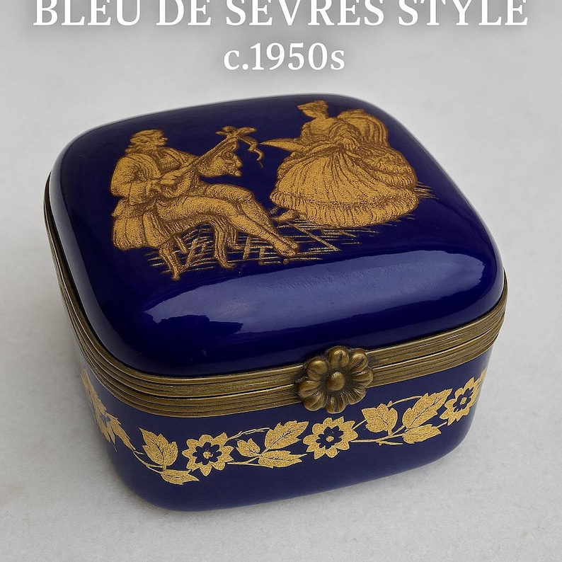 Puede incluir: Una caja de baratijas cuadrada azul cobalto con un dise&ntilde;o floral dorado y una tapa con bisagras. La tapa presenta una ilustraci&oacute;n dorada de dos figuras. El texto "BLEU DE SEVRES STYLE c.1950s" est&aacute; en la parte superior.