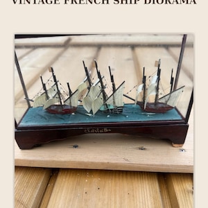 Op de afbeelding: Vintage Frans scheepsdiorama in een glazen kist. Het diorama toont drie miniatuurzeilschepen met witte zeilen en rode rompen, tegen een blauwe achtergrond. De basis is van donker hout, met de tekst "Chatelaillon - Atlantic Coast".