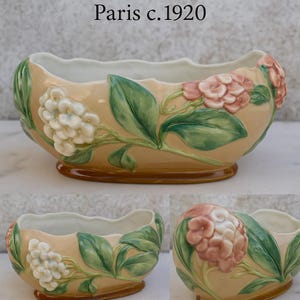 May include: A ceramic planter with a beige exterior and a scalloped white interior. The planter is adorned with raised floral and leaf designs in white, pink, and green. The text "Paris c.1920" is at the top.