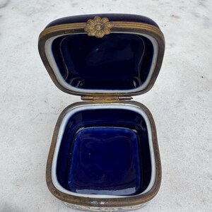 Puede incluir: Caja de baratijas azul cobalto antigua y abierta con marco de metal dorado. La caja tiene un cierre floral y un borde interior blanco. El interior de la caja es de color azul oscuro.