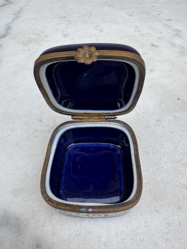 Puede incluir: Caja de baratijas azul cobalto antigua y abierta con marco de metal dorado. La caja tiene un cierre floral y un borde interior blanco. El interior de la caja es de color azul oscuro.