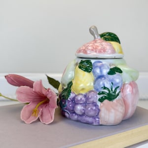 May include: A colorful ceramic jar with a lid, decorated with various fruits like grapes, pears, and peaches. The jar is placed on a stack of books, with a pink flower in the foreground. The jar is a decorative item.