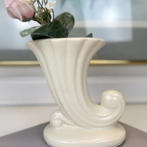 May include: Ivory-colored ceramic vase in a horn shape, with a curled base and fluted design. The vase holds a pink rose and small white flowers with green leaves. A decorative piece for floral arrangements.