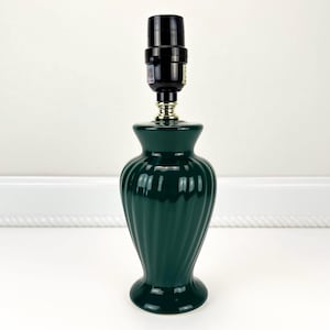 May include: A dark green ceramic table lamp with a black lamp socket. The lamp has a ribbed, vase-like base and a gold-colored accent at the neck. The lamp is designed for indoor use and is a decorative lighting fixture.