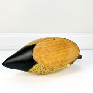 May include: A wooden duck figurine with a black beak and gold-colored accents. The duck's body is painted with a gold scale pattern. The underside of the duck is made of natural wood. A small brass-colored piece is attached to the bottom.