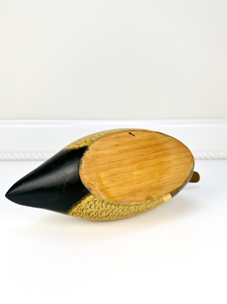 May include: A wooden duck figurine with a black beak and gold-colored accents. The duck's body is painted with a gold scale pattern. The underside of the duck is made of natural wood. A small brass-colored piece is attached to the bottom.