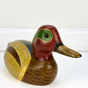 May include: A hand-painted wooden duck figurine. The duck features a red head with a green eye patch, a brown body with intricate detailing, and a yellow and blue wing. The beak is a light brown color.