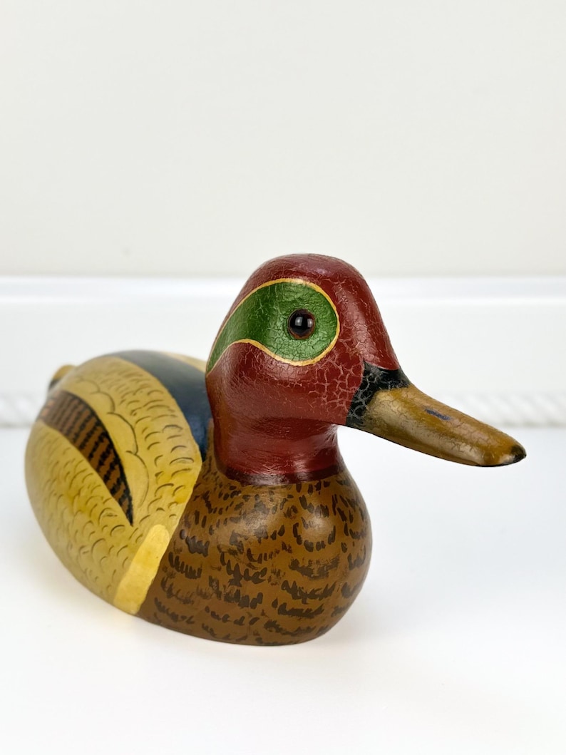 May include: A hand-painted wooden duck figurine. The duck features a red head with a green eye patch, a brown body with intricate detailing, and a yellow and blue wing. The beak is a light brown color.