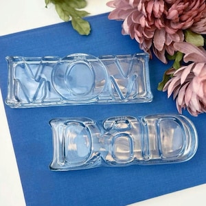 May include: Two light blue glass trays, one spelling "MOM" and the other "DAD". The trays are rectangular with the letters molded into the glass. The background is a deep blue surface with pink flowers.