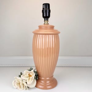 May include: A peach-colored ceramic lamp with vertical ribbed detailing. The lamp has a rounded base and a matching top. A black lamp socket and gold-toned hardware are at the top. A small bouquet of white and cream-colored flowers rests near the base of the lamp.