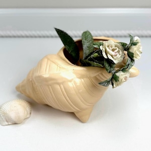 May include: A cream-colored ceramic seashell planter with a small seashell next to it. The planter is filled with green leaves and small white roses with pink centers. The planter has a textured surface and is resting on a white surface.