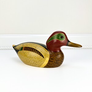 May include: A hand-painted wooden duck figurine. The duck features a red head with a green stripe, a brown body, and yellow and black wings. The beak is a light brown color. This decorative item is a charming addition to any home decor.