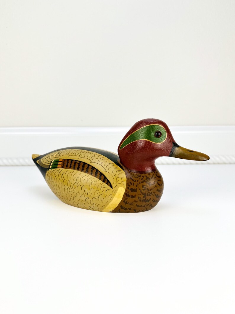 May include: A hand-painted wooden duck figurine. The duck features a red head with a green stripe, a brown body, and yellow and black wings. The beak is a light brown color. This decorative item is a charming addition to any home decor.