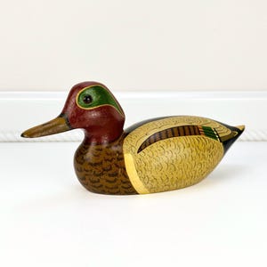 May include: A hand-painted wooden duck figurine. The duck features a burgundy head with a green accent, a brown beak, and a yellow and brown body. The duck is a decorative item.