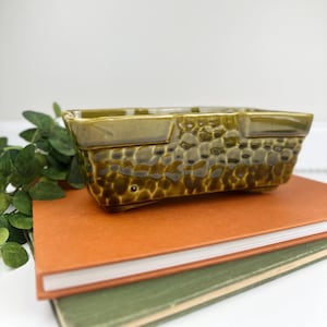 May include: A rectangular, olive green ceramic planter with a textured, dimpled surface. The planter has a raised rim and sits on small feet. The planter is displayed on top of an orange and green book.