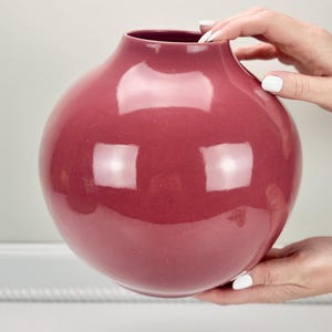May include: A glossy, round vase in a deep rose color. The vase has a wide, bulbous body and a narrow neck. The surface reflects light, creating highlights. The vase is held by two hands with white nail polish.