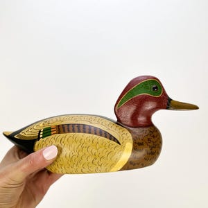May include: A hand-painted wooden duck figurine. The duck features a maroon head with a green stripe, a yellow and brown body, and a black tail. The beak is a light brown color. The figurine is held by a person.