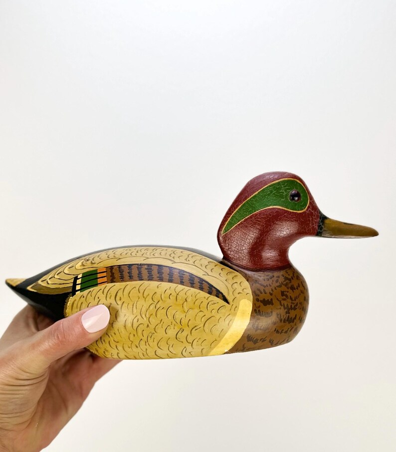 May include: A hand-painted wooden duck figurine. The duck features a maroon head with a green stripe, a yellow and brown body, and a black tail. The beak is a light brown color. The figurine is held by a person.