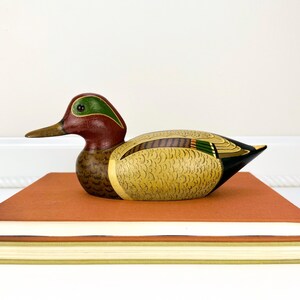 May include: A hand-painted wooden duck figurine with a red head, green accents, and a yellow and brown body. The duck is perched on a stack of books with orange and white covers. The duck's beak is a light brown color.