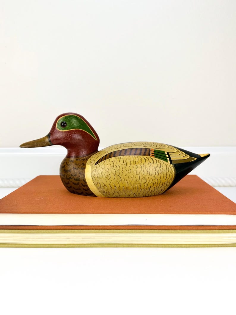 May include: A hand-painted wooden duck figurine with a red head, green accents, and a yellow and brown body. The duck is perched on a stack of books with orange and white covers. The duck's beak is a light brown color.
