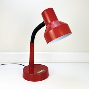 May include: A red desk lamp with a flexible black neck and a round base. The lamp has a conical shade with a white interior. The base has a small switch and a power cord.