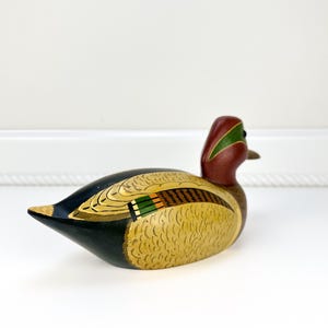 May include: A hand-painted wooden duck figurine. The duck features a red head with a green stripe, a yellow body with detailed feather patterns, and black and green wings. The duck is a decorative item.