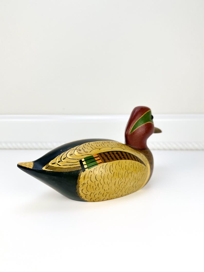 May include: A hand-painted wooden duck figurine. The duck features a red head with a green stripe, a yellow body with detailed feather patterns, and black and green wings. The duck is a decorative item.