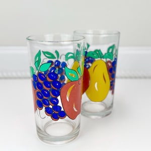 May include: Two clear drinking glasses with colorful fruit designs. One glass features blue grapes and a red apple, while the other has yellow pears and more grapes. The fruit illustrations are outlined in black and have green leaves.