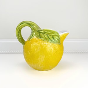 May include: A ceramic pitcher shaped like a lemon, with a green handle and a green leaf accent. The pitcher is yellow with a textured surface and a white rim.