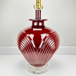 May include: A burgundy-colored table lamp with a ribbed, rounded base and a brass-colored lamp holder. The lamp sits on a clear, circular base. The lamp's design features vertical grooves that create a textured surface.