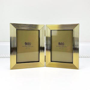 May include: A hinged, gold-colored photo frame with a ribbed design. The frame is made of solid brass and holds two 3.5" x 5" photos. The frame is displayed on a white surface.