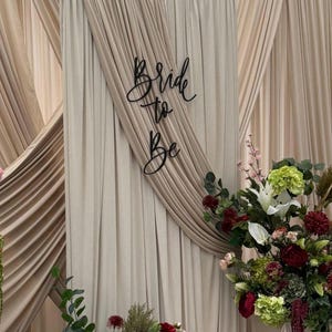 May include: A wedding or bridal shower backdrop with beige and tan draped fabric. The words "Bride to Be" are in black script. Floral arrangements with red, white, and green flowers are at the base of the backdrop, creating a decorative display.