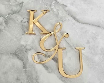 May include: A gold-toned reflective acrylic letter K, an ampersand symbol, and the letter U. The letters are arranged in a cluster with the K positioned above the ampersand and the U, which are slightly overlapping in a decorative arrangement.