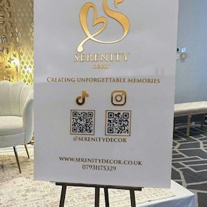 May include: A white sign with gold lettering and a gold logo, displayed on a wooden easel. The sign reads "SERENITY decor" and "Creating Unforgettable Memories." It includes social media icons and QR codes.