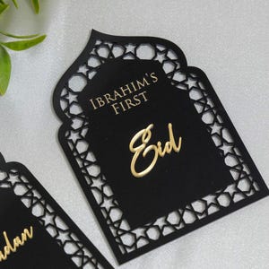May include: Black laser-cut card with a decorative geometric border. The card features the gold text "IBRAHIM'S FIRST" and "Eid". The design is in the shape of a mosque door, suitable for a holiday celebration.