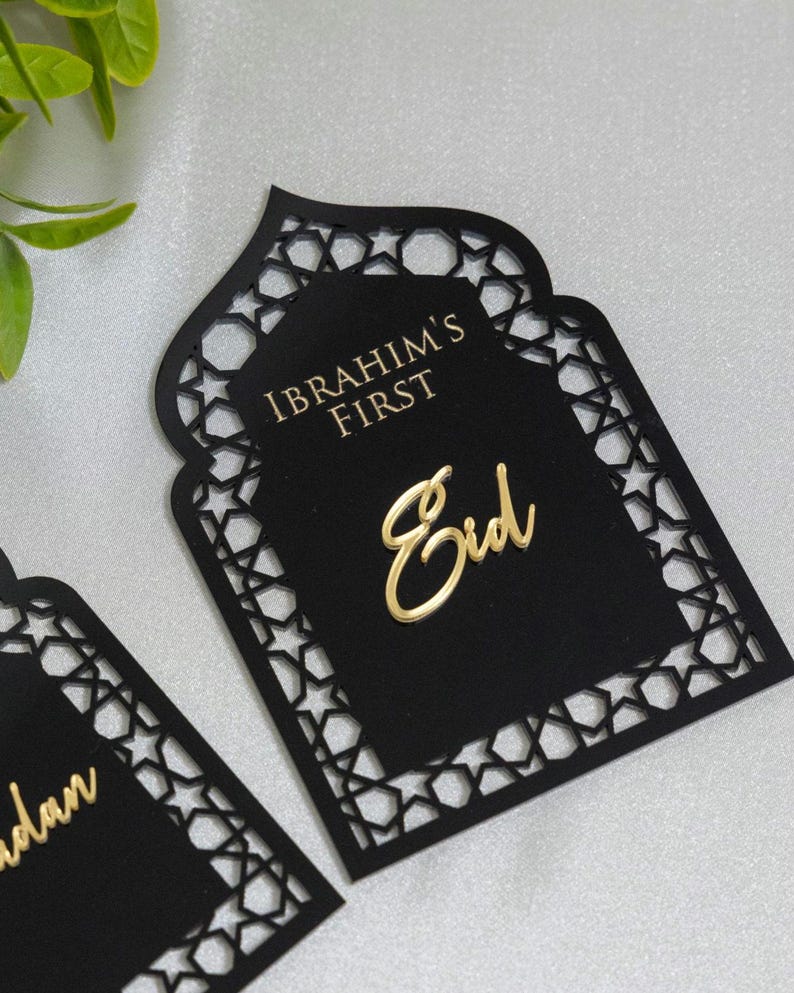May include: Black laser-cut card with a decorative geometric border. The card features the gold text "IBRAHIM'S FIRST" and "Eid". The design is in the shape of a mosque door, suitable for a holiday celebration.