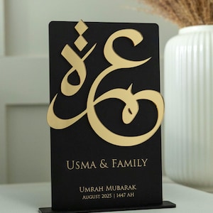 May include: A black rectangular plaque with gold Arabic calligraphy and the words "USMA & FAMILY". Below, it reads "UMRAH MUBARAK AUGUST 2025 | 1447 AH". The plaque is mounted on a small black stand.