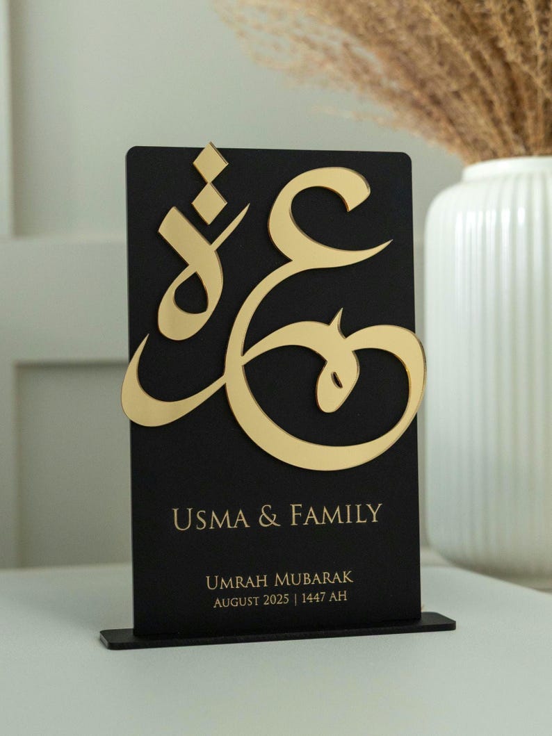 May include: A black rectangular plaque with gold Arabic calligraphy and the words "USMA & FAMILY". Below, it reads "UMRAH MUBARAK AUGUST 2025 | 1447 AH". The plaque is mounted on a small black stand.