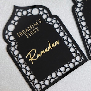 May include: Black laser-cut Ramadan decoration with a decorative star pattern border. The center features the text "IBRAHIM'S FIRST" and "Ramadan" in gold lettering. The decoration is in a unique arch shape.